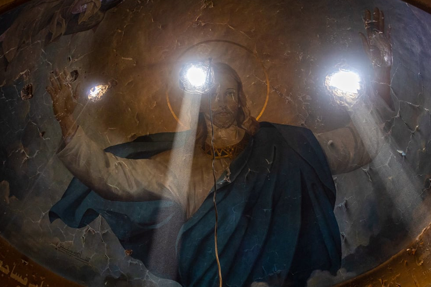 Light streams through three holes made by mortar rounds in the roof of Marmina Coptic Church, illuminating a mural of Jesus Christ.